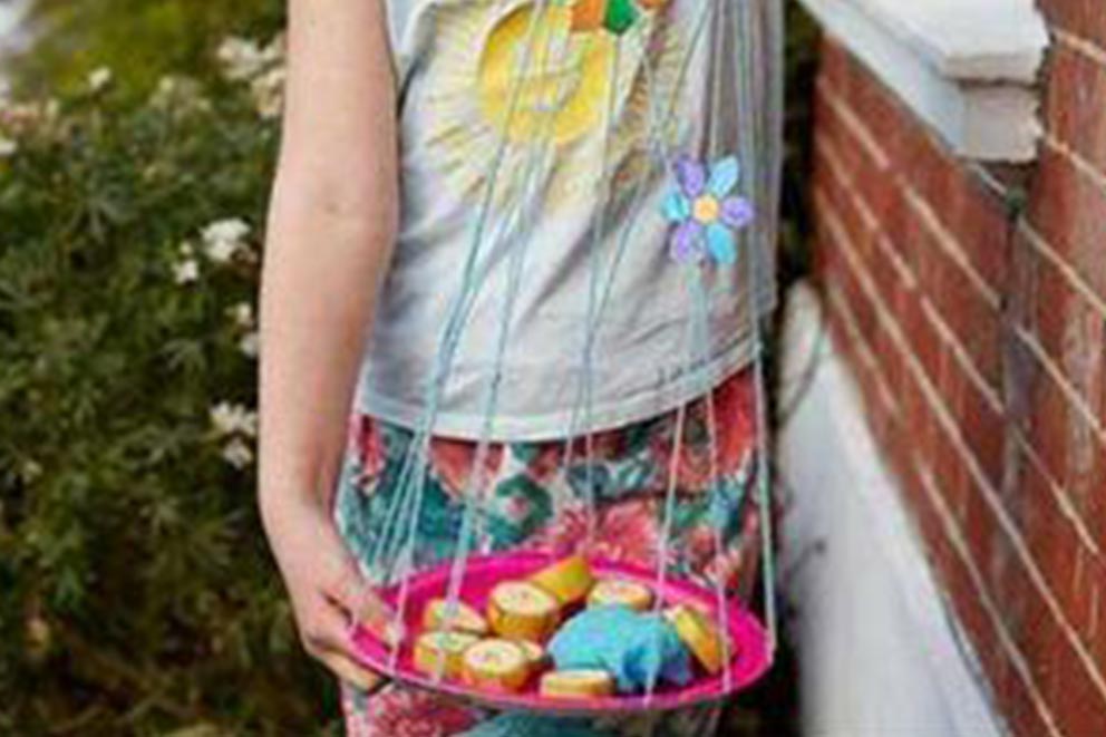 Child holding a homemade butterfly feeder made from a pink plate suspended by strings, decorated with flower cut-outs and filled with banana slices and a sugar-soaked cloth.