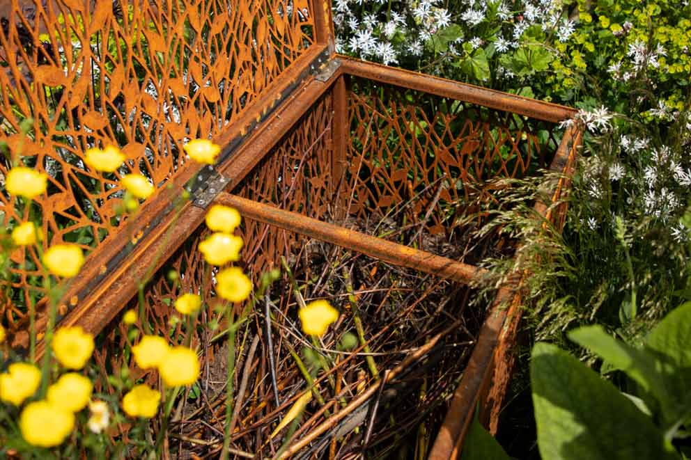 Rust-effect steel garden structure filled with twigs and branches, forming a tidy dead hedge surrounded by wildflowers.