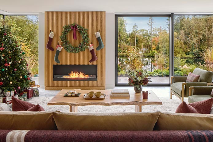 A large living space with feature fireplace, coffee table and corner sofa. The view looks out onto a lake.