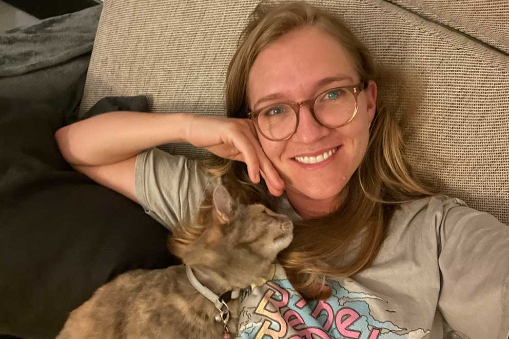 A smiling woman wearing glasses takes a selfie while relaxing on a sofa with her tabby cat resting on her chest.
