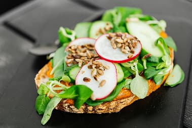 A slice of crusty bread is topped with an orange spread, fresh green leaves, sliced radish, cucumber and seeds.